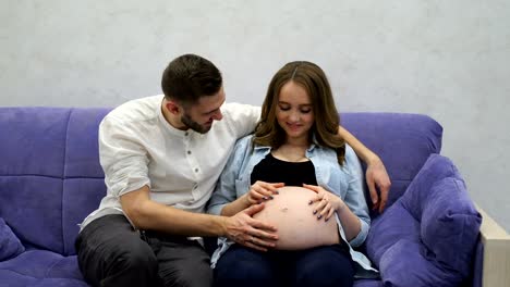 Schwangere-Frau-mit-ihrem-Mann-vor-dem-Fernseher-zu-Hause-auf-der-Couch-sitzen.-Ein-Mann-kommuniziert-mit-seiner-Frau-und-Kind.-Küssen-Sie-den-Bauch-zu-und-hören-Sie-die-Bewegungen-des-Babys-im-Bauch