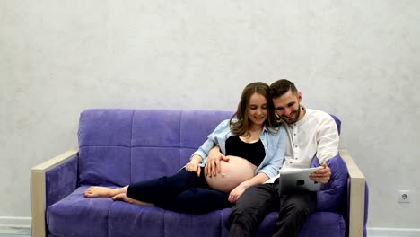 Happy-family-couple-sitting-on-the-couch-calling-on-video-communication-to-their-parents-in-another-city.-Tells-news-about-pregnancy.-A-pregnant-woman-and-a-man-communicate-via-a-tablet-computer-via-video-communication.-Video-conference.-Life-style