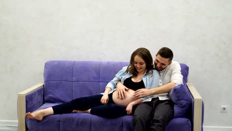 Happy-family-couple-sitting-on-sofa-watching-TV.-A-pregnant-woman-laughs-with-her-husband-while-watching-a-TV-show
