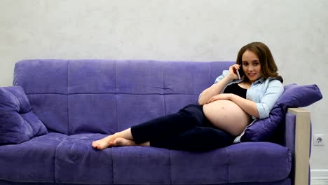 A-beautiful-pregnant-woman-sitting-on-the-couch-in-the-living-room-talking-on-a-mobile-phone-and-touching-her-stomach-with-her-hands-smiling.-Calls-her-husband