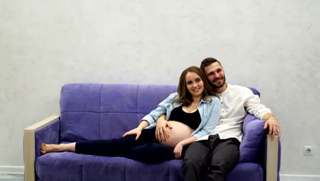 A-married-couple-is-sitting-at-home-on-the-couch-waiting-for-the-birth-of-a-child.-The-man-stroked-her-hand-across-her-belly-and-kissing-the-belly-of-his-pregnant-wife