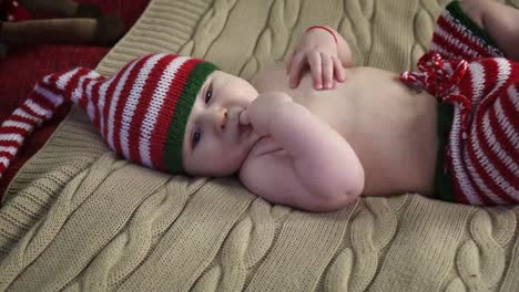 Peaceful-baby-boy-dressed-in-striped-cap-and-pants-is-lying-on-bed,-sucking-a-finger-and-smiling
