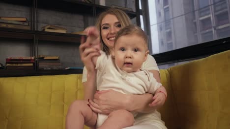 Happy-blonde-mother-and-her-son-are-sitting-on-yellow-couch,-looking-in-camera,-waving-and-smiling