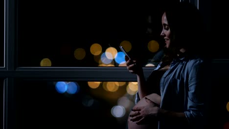 Pregnant-woman-at-night-talking-on-a-mobile-phone-touching-his-stomach-standing-at-the-large-panoramic-window-overlooking-the-city.-Cars-drive-by-outside-the-window.-The-girl-looks-at-the-phone-screen.-beautiful-bokeh-in-the-background