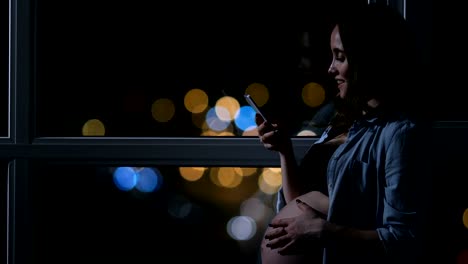 Mujer-embarazada-en-la-noche-hablando-en-un-teléfono-móvil,-tocando-su-estómago-de-pie-en-la-gran-ventana-panorámica-con-vistas-a-la-ciudad.-Coche-coches-por-fuera-de-la-ventana.-La-chica-se-ve-en-la-pantalla-del-teléfono.-hermoso-bokeh-en-el-fondo