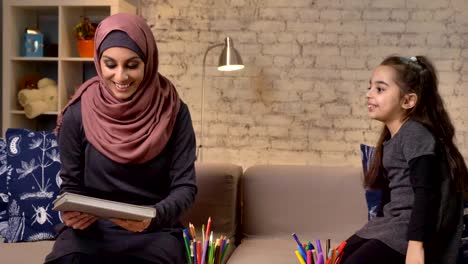 Young-Muslim-mother-in-a-hijab-takes-a-book-in-her-hands,-her-daughter-enthusiastically-laughs-and-claps-her-hands-50-fps