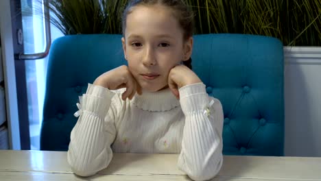 A-little-girl-with-freckles-on-her-face-in-a-white-dress-sitting-at-a-table-in-a-cafe-with-her-head-on-her-hands,-looking-at-the-camera-and-smiling.-She's-waiting-for-the-ordered-food.-Portrait.