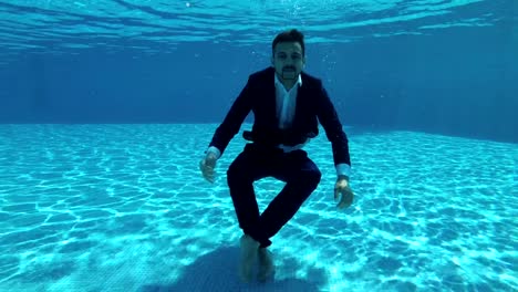 Asian-man-in-a-suit-and-a-white-shirt-dives-into-the-water-in-the-pool.-He-looks-at-the-camera-and-waves-his-hands.-Shooting-action-camera-under-water.