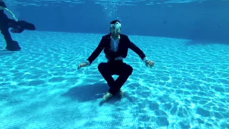 A-man-businessman-in-a-suit-and-a-white-shirt-dives-to-the-bottom-of-the-pool-under-the-water.-He-sits-on-the-bottom-in-a-Lotus-position,-looks-into-the-camera-and-lets-out-bubbles.-Slow-motion.-Action-camera-underwater.