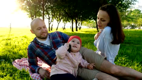 Bebé-niña-hablando-por-teléfono,-con-un-smartphone-en-sus-manos.-Familia-feliz-teniendo-un-picnic-al-aire-libre-en-verano-al-atardecer