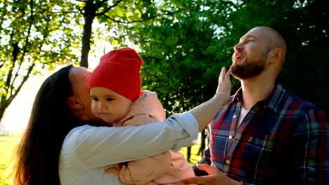 Glückliche-Eltern-spielen-mit-einem-Kind-in-einer-Nahaufnahme-Park.-Junge-Familie-mit-Baby-umarmen-und-küssen-einander-im-Sommerpark-bei-Sonnenuntergang