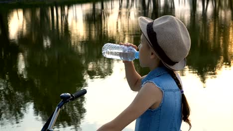 Glückliche-kleine-Mädchen-mit-dem-Fahrrad-in-die-Kamera-schaut-und-den-Sonnenuntergang-auf-dem-River-Bank-lächelte.-Sie-steht-in-einem-Hut-auf-einem-Hintergrund-von-Wasser-und-trinkt-Wasser-aus-einer-klaren-Flasche.