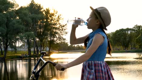 Schöne-kleine-Mädchen-Trinkwasser-aus-einer-transparenten-Flasche-bei-Sonnenuntergang.-Sie-steht-in-einem-Hut-mit-einem-Fahrrad-auf-dem-River-Bank-in-der-Sonne.