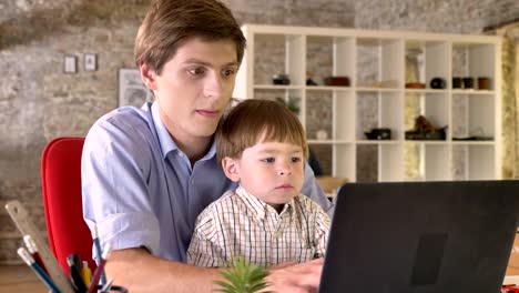 Young-father-holding-his-little-son-and-working-on-laptop,-busy,-sitting-in-modern-office