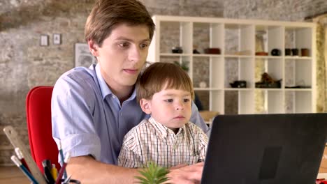 Joven-padre-sosteniendo-a-su-pequeño-hijo-y-trabajando-en-ordenador-portátil,-ocupado,-sentado-en-la-oficina-moderna