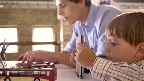 Pequeño-hijo-mirando-tablet-cuando-su-padre-joven-trabajando-en-la-computadora-portátil,-sentado-en-la-oficina-de-ladrillo-moderno