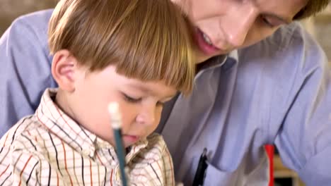 Joven-padre-y-su-pequeño-hijo-pintura-sobre-papel-y-explotación-tableta,-papá-ayudando-y-enseñando-a-su-hijo-a-escribir