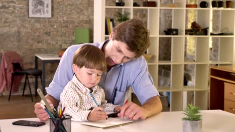 Young-caucasian-father-holding-his-son-and-teaching-him-how-to-write,-sitting-in-modern-office