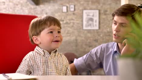 Young-business-father-twirling-around-his-little-son-on-chair-and-talking-on-phone-in-modern-office