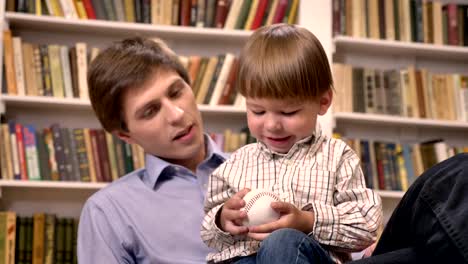 Little-boy-sitting-with-his-young-father-and-holding-ball,-shelves-with-books-background