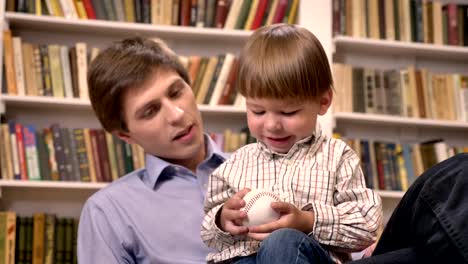 Kleiner-Junge-sitzt-mit-seinem-jungen-Vater-und-Holding-Ball,-Regale-mit-Büchern-Hintergrund