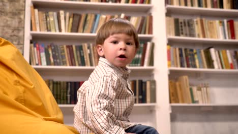 Little-cute-kid-sitting-on-chair-and-catching-ball-from-somebody,-bookshelves-background