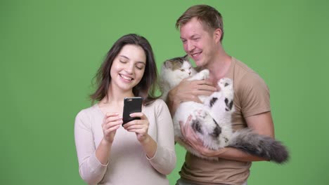 Young-couple-using-phone-together-with-cat-against-green-background