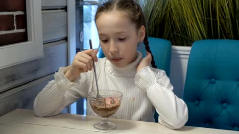 Linda-niña-con-coletas-sentada-y-comiendo-helado-de-fresa-y-chocolate-en-el-café.-Ella-come-con-una-cuchara-larga-de-cristal-taza.-Retrato