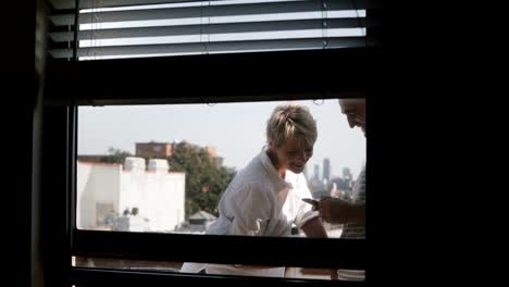 Happy-smiling-romantic-couple-standing-at-a-small-sunny-balcony,-view-through-apartment-window,-using-smartphones