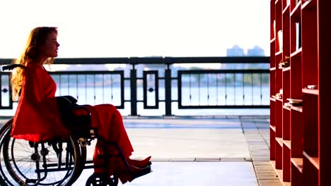 Young-Red-Haired-Girl-With-A-Disability-In-Orange-Dress-Pulls-Up-On-A-Wheelchair-To-The-Bookshelf-And-Takes-The-Book