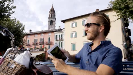 Young-man-using-digital-tablet-in-Swiss-village-near-lake-and-mountains--sitting-on-bench-with-bicycle-beside-him.-People-travel-technology-portable-devices-concept