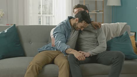 Cute-Attractive-Male-Gay-Couple-Sit-Together-on-a-Sofa-at-Home.-Boyfriend-Puts-His-Hand-on-Partner's-and-They-Hug.-They-are-Happy-and-Smiling.-They-are-Casually-Dressed-and-Room-Has-Modern-Interior.