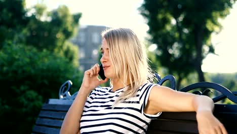 Attractive-blonde-young-woman-sitting-in-park-talking-on-her-mobile-phone