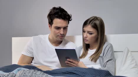 Relaxing-Young-Couple-Browsing-Internet-on-Tablet-in-Bed