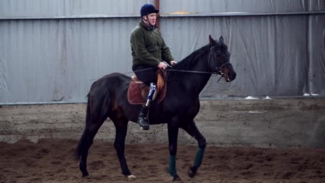 in-special-hangar,-a-young-disabled-man-learns-to-ride-a-black,-thoroughbred-horse,-hippotherapy.-man-has-an-artificial-limb-instead-of-his-right-leg.-concept-of-rehabilitation-of-disabled-with-animals