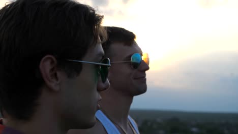 Profile-of-young-male-couple-in-sunglasses-standing-on-the-edge-of-rooftop-and-talking.-Handsome-gay-boys-relaxing-on-roof-of-high-rise-building-and-enjoying-beautiful-cityscape.-Close-up-Slow-motion