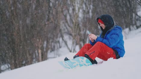 A-male-snowboarder-sitting-on-the-snow-takes-photos-on-the-phone-of-a-beautiful-resort-landscape-for-social-networks.-Blogger-at-the-resort.-Write-text-messages-to-friends-using-your-smartphone