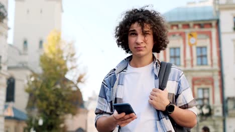 Young-curly-man,-tourist-or-student,-generation-z-in-hipster-outfit,-walking-through-the-street,-using-his-smartphone-scrolls-through-social-media-feed-on-device,-checking-map-or-reading-news-on-app