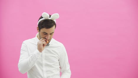 Young-handsome-boy-standing-on-a-pink-background.-A-man-with-rabble-ears-on-his-head.-A-man-holding-a-carrot-puts-it-in-his-mouth-and-reproduces-the-movements-of-a-cigarette-with-a-cigarette-lighter.