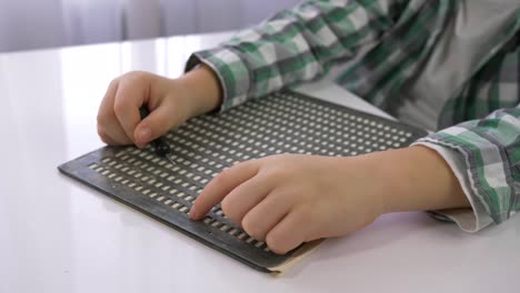 Niño-con-discapacidades-visuales-aprendiendo-a-escribir-caracteres-Braille-de-fuente-sentado-en-la-mesa-en-la-habitación-luminosa,-las-manos