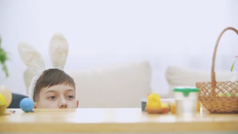 Cute-little-boy-with-bunny-ears-is-hiding-under-the-table-full-of-Easter-decorations.-Little-cute-white-bunny-is-attacking-boy-kindly.-Laught-is-in-the-room.