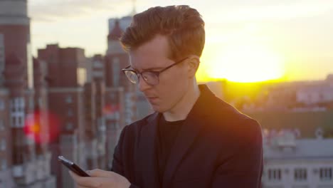 Stylischer-Mann-mit-Smartphone-auf-Rooftop-bei-Sonnenuntergang