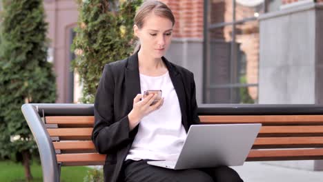 Young-Businesswoman-Using-Smartphone-and-Laptop,-Sitting-on-Bench