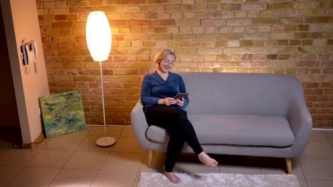 Closeup-shoot-of-senior-caucasian-woman-texting-on-the-tablet-and-laughing-cheerfully-sitting-on-the-couch-in-a-cozy-apartment