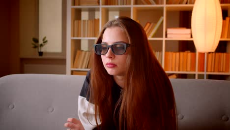 Portrait-of-young-teenage-girl-in-3d-glasses-watches-film-on-TV-sitting-on-sofa-on-bookshelves-background-at-home.