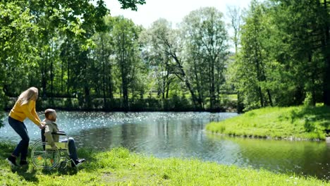 Caring-senior-woman-pushing-her-disabled-husband-in-wheelchair-towards-lake-in-green-park.-Couple-looking-at-scenic-view-and-talking