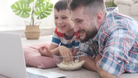 Padre-con-hijo-comiendo-palomitas-de-maíz-mientras-ve-película-divertida-en-el-ordenador-portátil