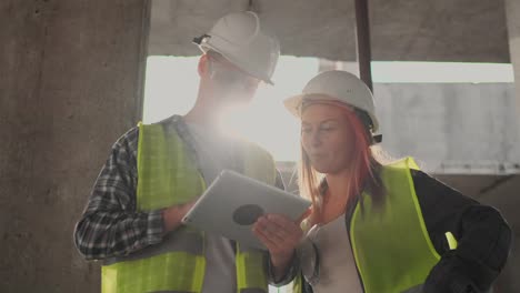 Trabajadorde-la-construcción-hombre-y-arquitecto-mujer-en-un-casco,-discutir-el-plan-de-construcción-de-la-casa,-hablarse-unos-a-otros-sobre-el-diseño,-sosteniendo-una-tableta,-mirar-los-dibujos,-fondo-de-los-rayos-del-sol