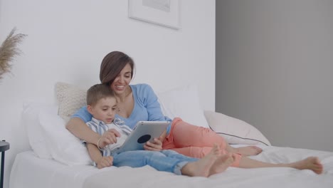 Mamá-e-hijo-mirando-la-pantalla-de-la-tableta-acostado-en-una-cama-blanca.-Juega-con-tu-hijo-en-tu-tablet-y-mira-videos-divertidos