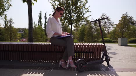 Joven-e-independiente-empresaria-en-un-banco-en-un-parque,-junto-a-una-moto-eléctrica.